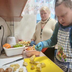 Dwie kobiety w kuchni przygotowują owoce. Jedna z nich, w niebieskich rękawicach, kroi banany na żółtej desce do krojenia, a druga obserwuje z zainteresowaniem. Na blacie znajdują się również inne owoce, takie jak kiwi, winogrona, pomarańcze i pokrojone jabłka.